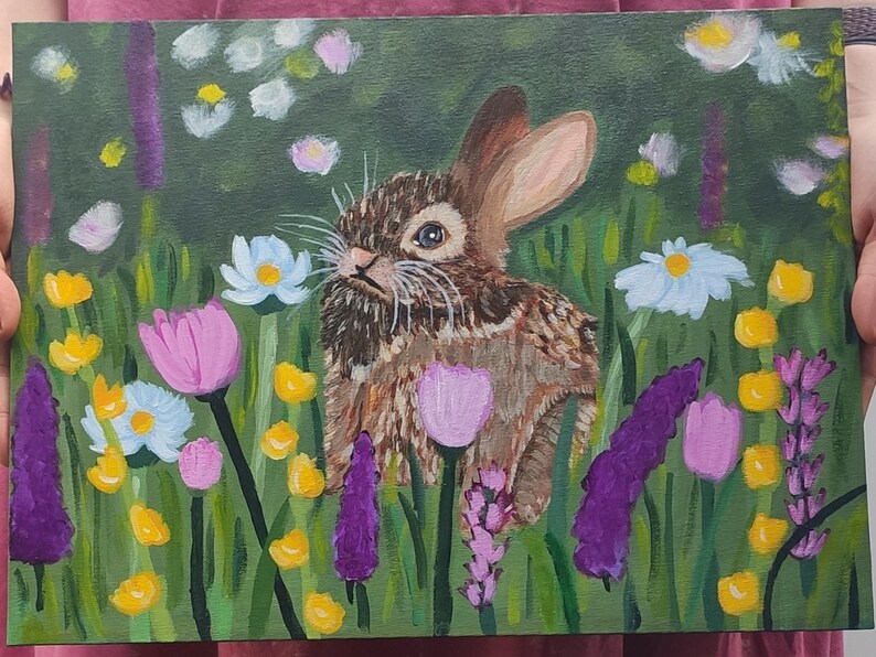 May include: An acrylic painting on canvas depicting a brown rabbit sitting in a field of colorful wildflowers. The scene features purple, pink, white, and yellow flowers set against a dark green background with visible brushstrokes throughout.