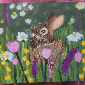 May include: An acrylic painting on canvas depicting a brown rabbit with long ears positioned in the center of a lush green meadow filled with colorful wildflowers including white daisies, yellow buttercups, and purple lavender stalks.