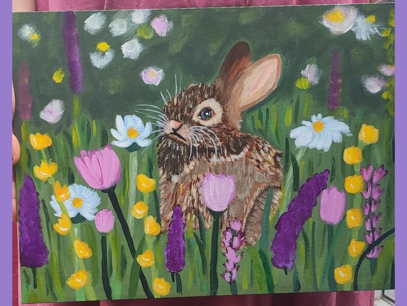 May include: An acrylic painting on canvas depicting a brown rabbit with long ears positioned in the center of a lush green meadow filled with colorful wildflowers including white daisies, yellow buttercups, and purple lavender stalks.