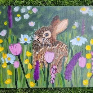 May include: An acrylic painting on canvas depicting a brown rabbit sitting in a field of colorful wildflowers. The scene features purple, pink, yellow, and white flowers set against a green background, with the rabbit positioned in the center.
