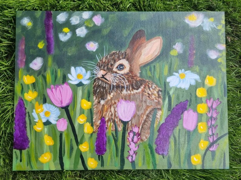 May include: An acrylic painting on canvas depicting a brown rabbit sitting in a field of colorful wildflowers. The scene features purple, pink, yellow, and white flowers set against a green background, with the rabbit positioned in the center.