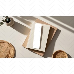 May include: A white rectangular bookmark on top of two beige books. The books are on a light beige surface with a woven basket and a potted plant in the background.