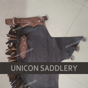 May include: Brown leather chaps with fringe and tooled leather accents. The chaps have a black suede body and are decorated with silver studs. The text "UNICON SADDLERY" appears on the chaps.