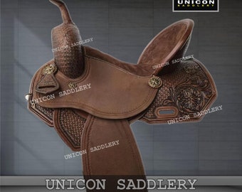 Könnte beinhalten: Ein brauner Ledersattel mit aufwendigen floralen Verzierungen. Der Sattel verfügt über einen hohen Hinterzwiesel und einen breiten Sitz. Der Sattel ist mit dem Text "UNICON SADDLERY" gebrannt.