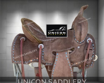 May include: A brown leather saddle with blue stitching and a tooled design. The saddle has a horn, stirrups, and a cinch. The saddle is labeled "UNICON SADDLERY".