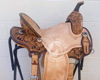 May include: A tan leather saddle with intricate floral tooling and a silver concho. The saddle is on a black metal stand and has a stirrup on the right side. The saddle is labeled "Unicon Saddlery".