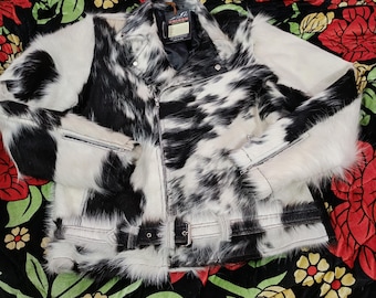 May include: A black and white cowhide jacket with a zipper closure, belt, and white fur trim. The jacket has a unique spotted pattern and a tag that reads "UNICORN".