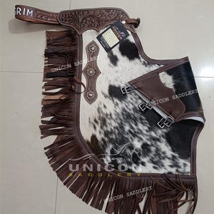 Op de afbeelding: Bruine leren chaps met franjes, met een wit en zwart koehuidpatroon. De chaps hebben een decoratief bloemenpatroon en metalen accenten. De tekst "UNICON SADDLERY" is zichtbaar op de chaps.