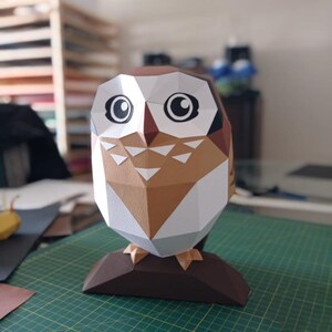 May include: A papercraft owl sculpture with brown, white and black details. The owl has large, round eyes and a triangular beak. The sculpture is standing on a brown base.