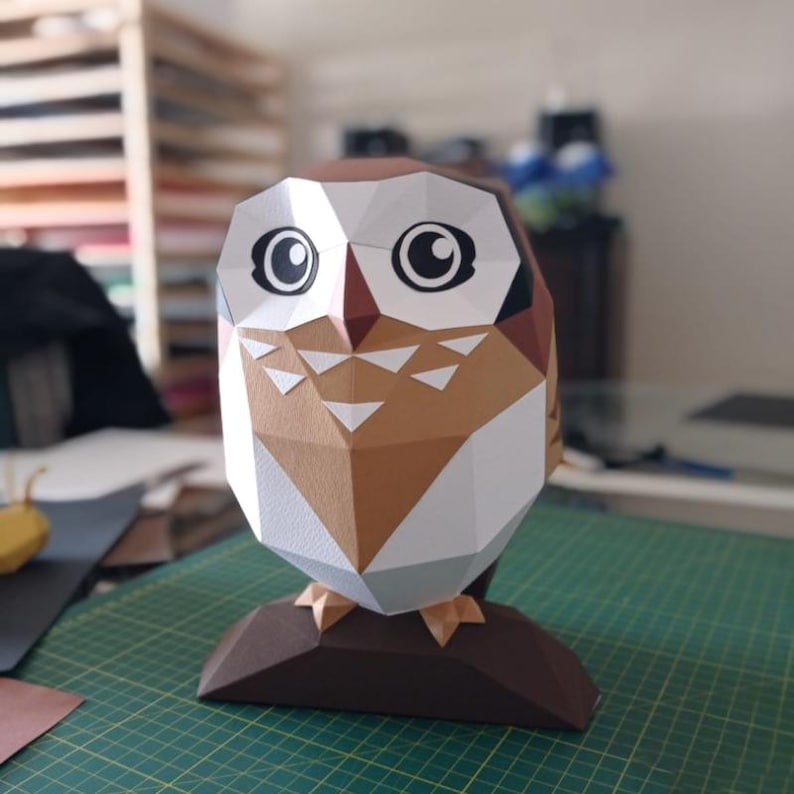 May include: A papercraft owl sculpture with brown, white and black details. The owl has large, round eyes and a triangular beak. The sculpture is standing on a brown base.