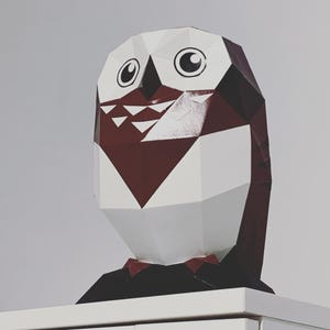 May include: A white and burgundy papercraft owl with black eyes and a black bow tie. The owl is sitting on a white surface.