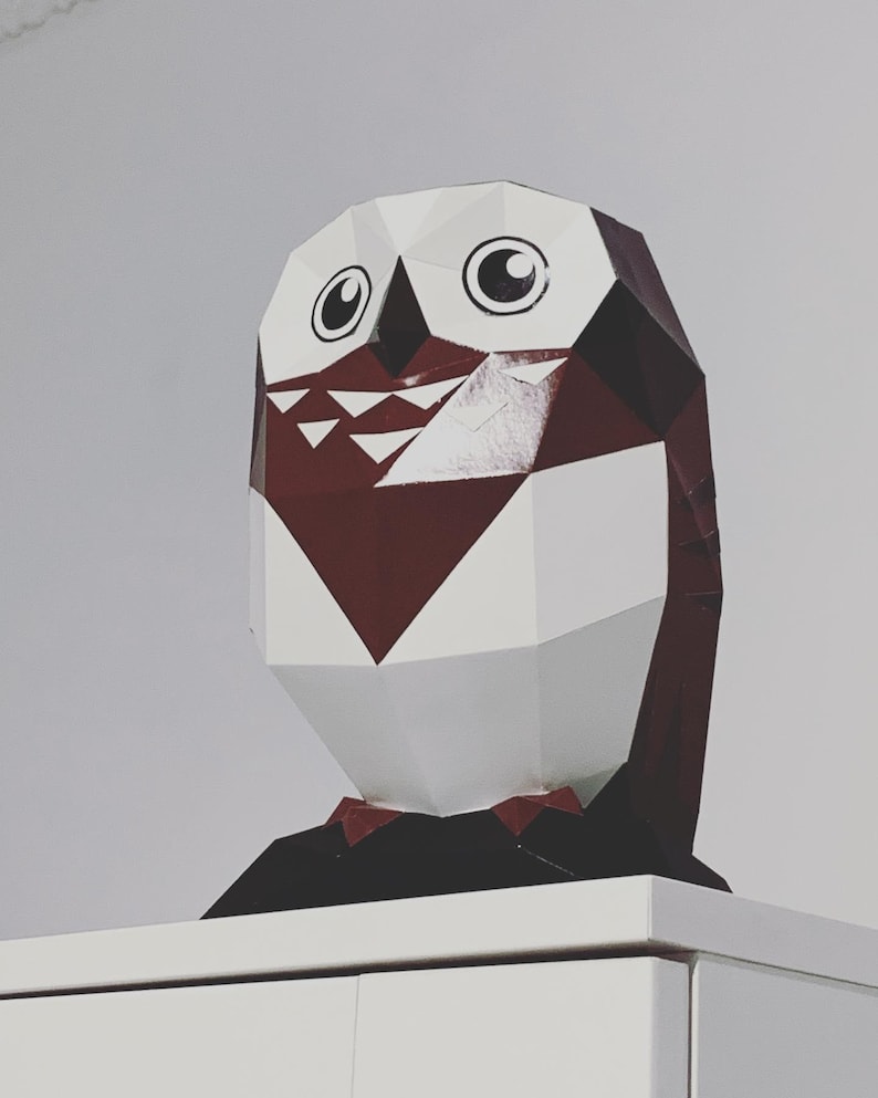 May include: A white and burgundy papercraft owl with black eyes and a black bow tie. The owl is sitting on a white surface.
