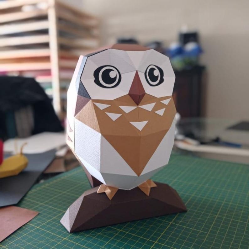 May include: A papercraft owl with brown, white and black accents. The owl has large eyes and a beak. The owl is standing on a brown base.