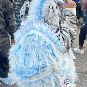 Op de afbeelding: Een lichtblauwe nep-bont rugzak met een witte rand en een zwart-witte tie-dye hoodie. De rugzak heeft een QR-code label eraan bevestigd.