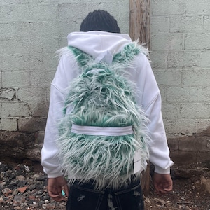 May include: A mint green and white faux fur backpack featuring a long-pile texture, a white horizontal zip across the front pocket, and a white rectangular tag attached to the lower right side, worn over a plain white hooded sweatshirt.