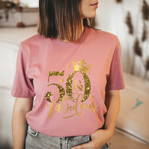Pode incluir: Uma camiseta rosa com um design de glitter dourado que diz "50 & fabuloso" com uma coroa acima do 50.