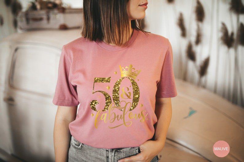 Pode incluir: Uma camiseta rosa com um design de glitter dourado que diz "50 & fabuloso" com uma coroa acima do 50.