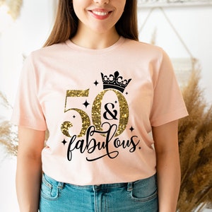 Pode incluir: Uma camiseta rosa com um design de glitter dourado que diz "50 & fabuloso" com uma coroa preta acima do 50. A camiseta est&aacute; em uma pessoa que est&aacute; usando jeans azul.