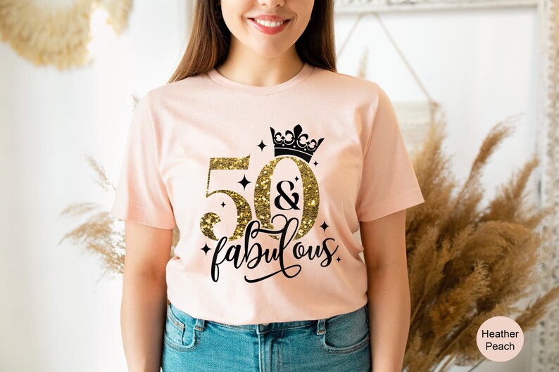 Pode incluir: Uma camiseta rosa com um design de glitter dourado que diz "50 & fabuloso" com uma coroa preta acima do 50. A camiseta est&aacute; em uma pessoa que est&aacute; usando jeans azul.