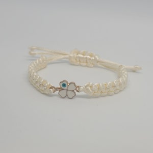 May include: White string bracelet with a gold four-leaf clover charm featuring a blue evil eye in the centre.