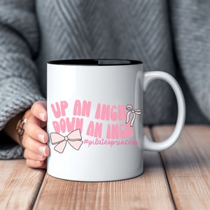 May include: White ceramic coffee mug with a black rim. The mug has a pink design that says "Up an inch down an inch #pilatesprincess".