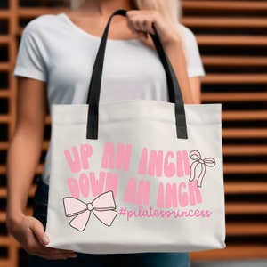May include: White canvas tote bag with black handles. The bag has pink text that reads "Up an inch down an inch #pilatesprincess" and a pink bow.