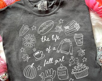 May include: Dark gray Comfort Colors t-shirt with white line art. The shirt features the text "the life of a fall girl" surrounded by drawings of fall-themed items like a pumpkin, coffee cup, and sweater.