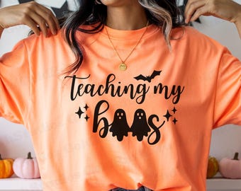 May include: Orange t-shirt with the phrase "Teaching my boos" in black script, featuring two ghost figures and a bat. The shirt is a Halloween-themed design.