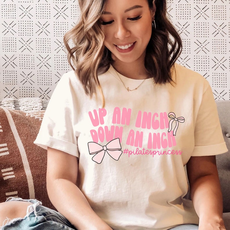 May include: White t-shirt with pink text that reads "Up an inch down an inch #pilatesprincess" and a pink bow graphic.