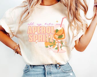 May include: A cream-coloured t-shirt featuring the text "off my tits on APEROL SPRITZ" in pink and orange. The design includes a cocktail glass with an orange slice, a straw, and floral details. The shirt is worn with blue denim jeans.