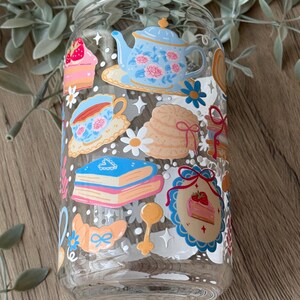 May include: Clear glass tumbler with colorful illustrations of a teapot, teacup, cake, and other tea party-themed graphics. The design includes pink, blue, and orange elements, creating a whimsical aesthetic. The tumbler is ideal for cold beverages.