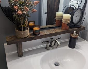 May include: A rustic wooden shelf spans a bathroom sink, holding candles, a clock, and a soap dispenser. A vase with floral arrangement sits on the left. The mirror reflects the items, creating a warm, inviting atmosphere.