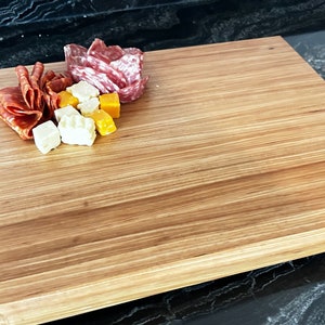 May include: A rectangular wooden chopping board with a natural wood grain finish. The board has a rounded edge and is made of light brown wood. There are small pieces of salami and cheese on the board.