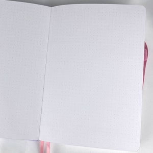 May include: A white notebook with a pink ribbon bookmark. The notebook has a dotted grid pattern on the pages.