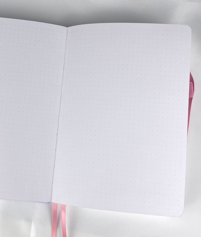 May include: A white notebook with a pink ribbon bookmark. The notebook has a dotted grid pattern on the pages.