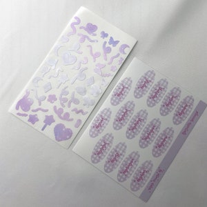 May include: Two sheets of stickers with a pastel purple and white color scheme. One sheet features a variety of shapes and designs, including stars, hearts, moons, and swirls. The other sheet features 16 oval stickers with a gingham pattern and the text "Happy" written in pink cursive.
