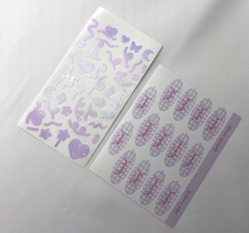 May include: Two sheets of stickers with a pastel purple and white color scheme. One sheet features a variety of shapes and designs, including stars, hearts, moons, and swirls. The other sheet features 16 oval stickers with a gingham pattern and the text "Happy" written in pink cursive.
