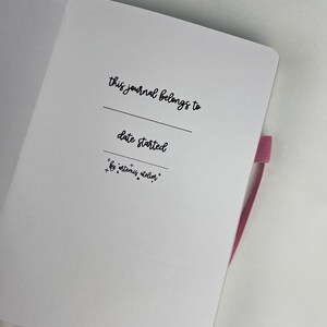 May include: A white journal with a black inscription that reads "this journal belongs to" and "date started". The inscription also includes the text "by artemis atelier".