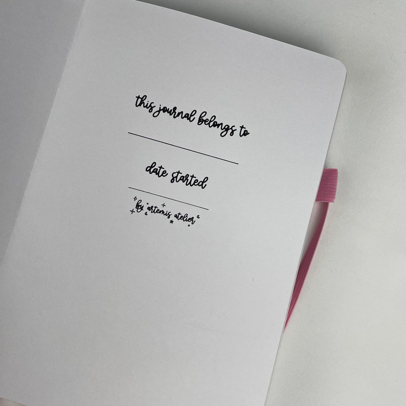 May include: A white journal with a black inscription that reads "this journal belongs to" and "date started". The inscription also includes the text "by artemis atelier".