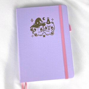May include: A lavender-colored notebook with a pink ribbon bookmark. The cover features a gold foil design of two cartoon bears wearing witch hats and holding wands.