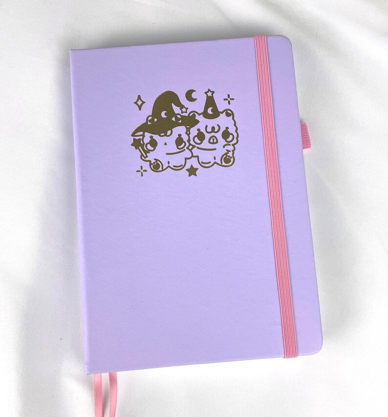 May include: A lavender-colored notebook with a pink ribbon bookmark. The cover features a gold foil design of two cartoon bears wearing witch hats and holding wands.