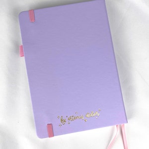May include: A lavender notebook with a pink ribbon bookmark and the text "by artemis atelier" in gold lettering.