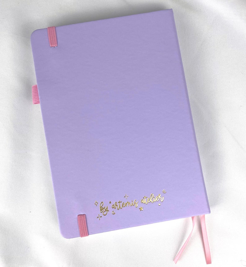 May include: A lavender notebook with a pink ribbon bookmark and the text "by artemis atelier" in gold lettering.