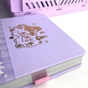 May include: A lavender-colored notebook with a gold foil design of two cartoon characters wearing witch hats. The notebook has a pink ribbon bookmark.