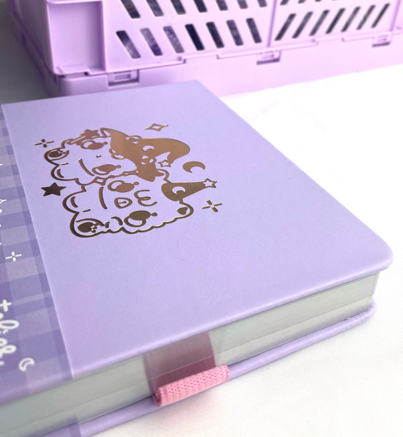 May include: A lavender-colored notebook with a gold foil design of two cartoon characters wearing witch hats. The notebook has a pink ribbon bookmark.