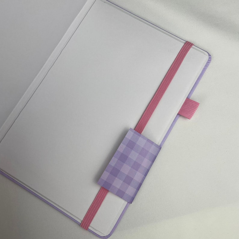 May include: A purple notebook with a pink elastic closure and a purple and white checkered bookmark.