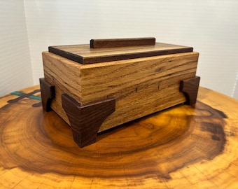 May include: A handcrafted wooden box with a rectangular shape and a lid. The box is made of light-colored wood with darker wood accents on the lid and corners. The box has a small handle on the lid and is displayed on a wooden surface.