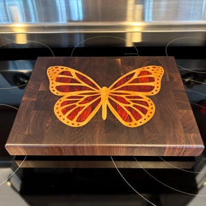 May include: A square, end-grain cutting board with a butterfly inlay. The butterfly design features orange and red hues, set against a dark brown wood background. The board is approximately 12 inches square, suitable for kitchen use.