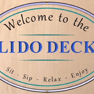 May include: A wooden sign with an oval shape and a blue and teal border. The sign says "Welcome to the Lido Deck" in black text and "Sit - Sip - Relax - Enjoy" in teal text.