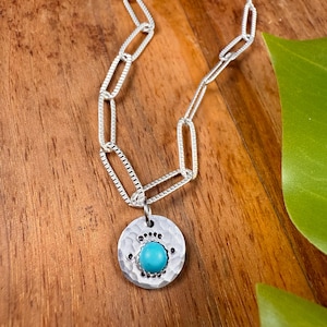 May include: A silver necklace with a hammered pendant featuring a turquoise stone and black accents. The pendant is round and hangs from a chain with rectangular links. The necklace rests on a wooden surface.